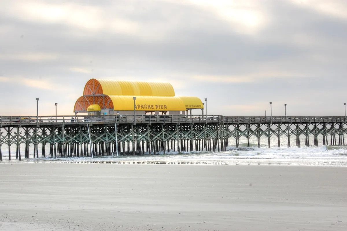 This exterior scene focuses on Apache Pier stretching into the ocean, with waves lapping below and a distinctive golden roof. The setting is lively and inviting, suggesting opportunities for beachside walks, fishing, photography, and group adventures.