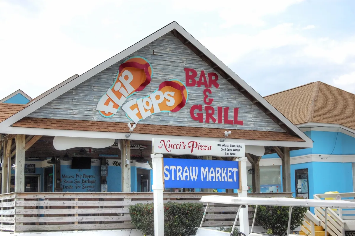 A vibrant strip of storefronts featuring signage for Flip Flops Bar & Grill, Nucci's Pizza, and Straw Market. The area is bustling with opportunities for dining, shopping, and socializing, reflecting the lively local scene.