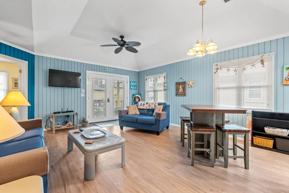 A cheerful living and dining space with blue walls, a cushioned sofa, a family-sized dining table, and a television set. The room’s layout encourages socializing, watching movies, and enjoying shared meals, while decorative touches and ample lighting create a relaxed ambiance.