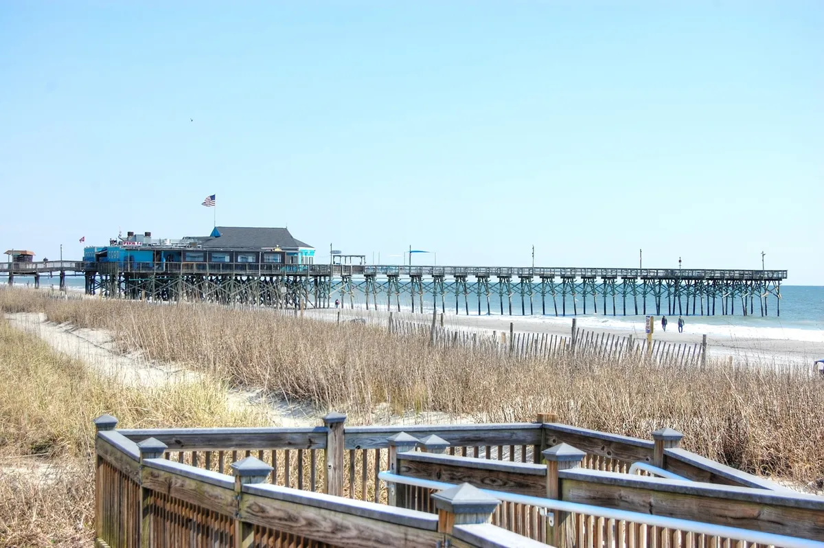 A serene stretch of sandy beach with gently rolling waves, dune grasses, a wooden pier extending into the blue sky, and open coastal views. The scene captures the calm and natural beauty found just a short walk from the community, echoing the promise of sunrise adventures and easy beach access.