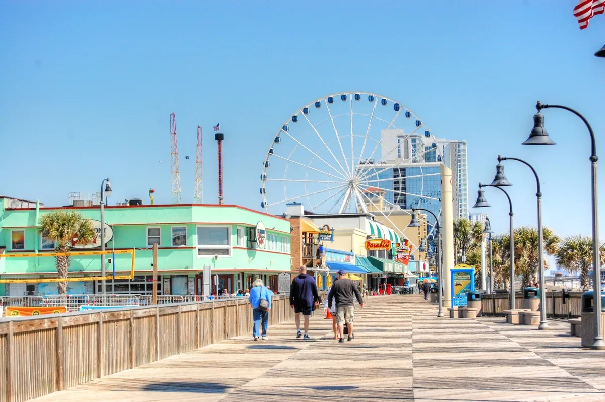 This lively exterior scene features the iconic Myrtle Beach Ferris wheel, colorful boardwalk buildings, and people enjoying a stroll. The setting is vibrant and energetic, capturing the excitement of local attractions.