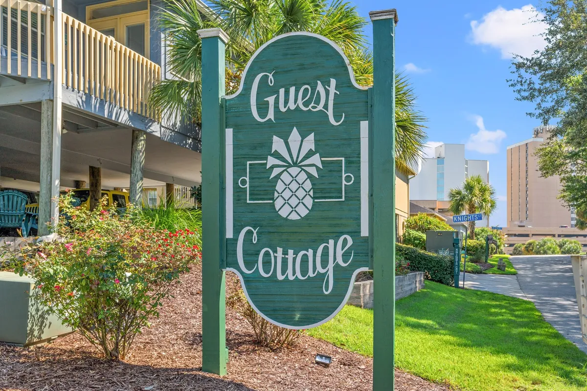 The image shows the exterior of a guest cottage, framed by palm trees and beautifully landscaped gardens. The prominent 'Guest Cottage' sign sets the scene, while the surrounding greenery and charming architecture convey a warm, welcoming atmosphere ideal for group gatherings.