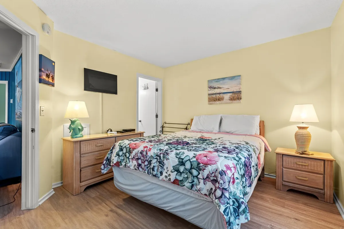 A cozy bedroom featuring a plush queen bed dressed in a floral blanket, with matching nightstands and elegant lamps on either side. The serene wall art and soft lighting contribute to the peaceful, inviting atmosphere, making it an ideal space for unwinding after a day at the beach.