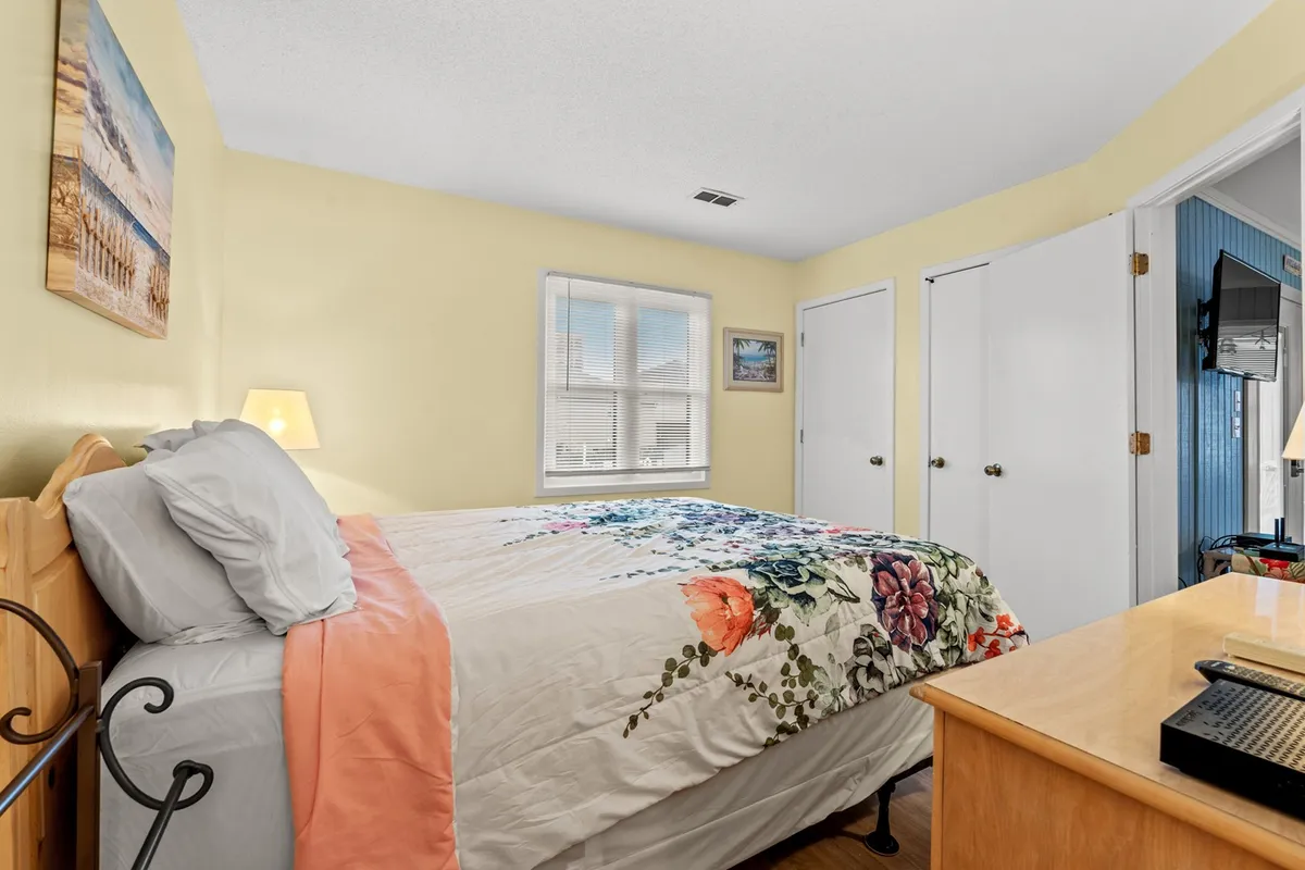 A tranquil bedroom highlighted by a queen bed with a floral bedspread, a wooden dresser for ample storage, and a window allowing natural light to stream in. Artwork on the wall and a peaceful ambiance make the room inviting for extended stays.
