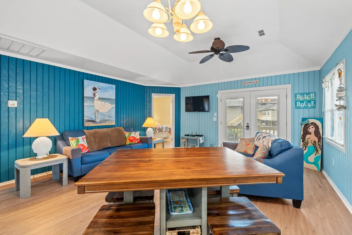 This image shows a wooden dining table surrounded by comfortable seating, set against blue walls with decorative artwork. Sunlight pours in through open doors, creating a light and airy feel perfect for gatherings and shared meals.